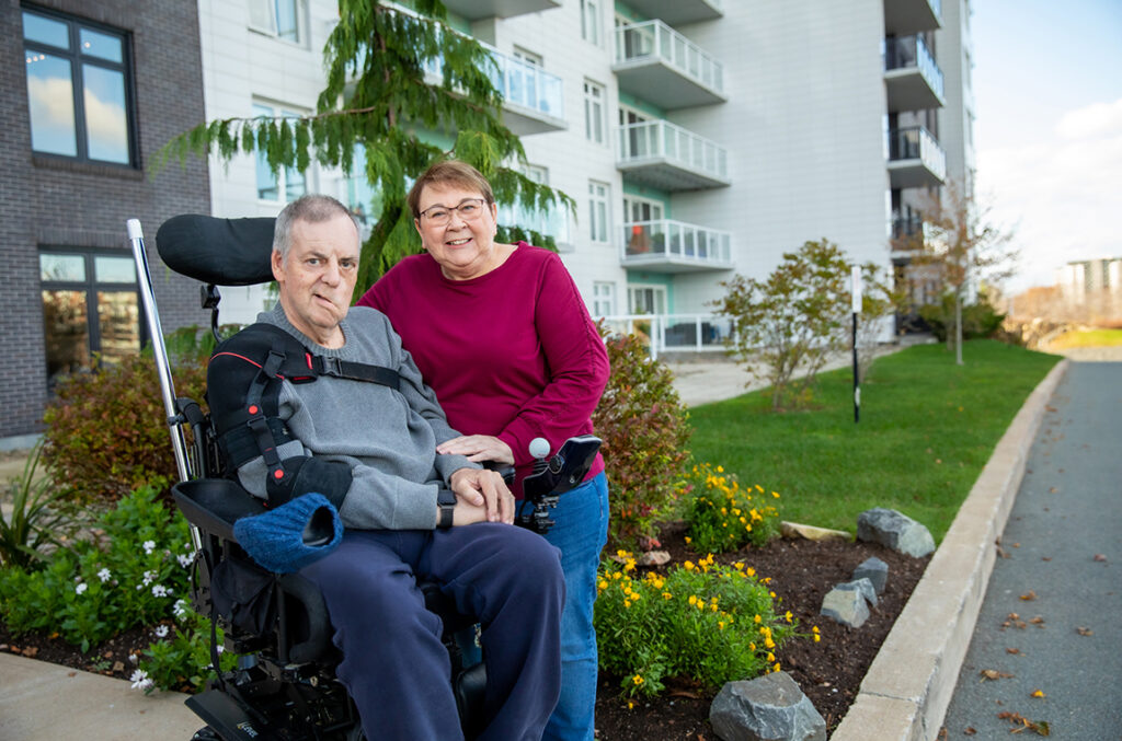 Aileen Nauss standing beside Charles Nauss in a wheelchair outside apartment building