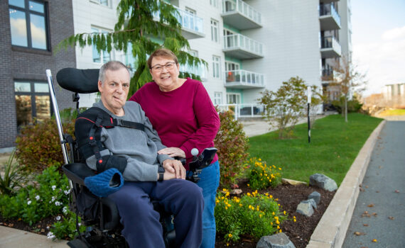 Aileen Nauss standing beside Charles Nauss in a wheelchair outside apartment building